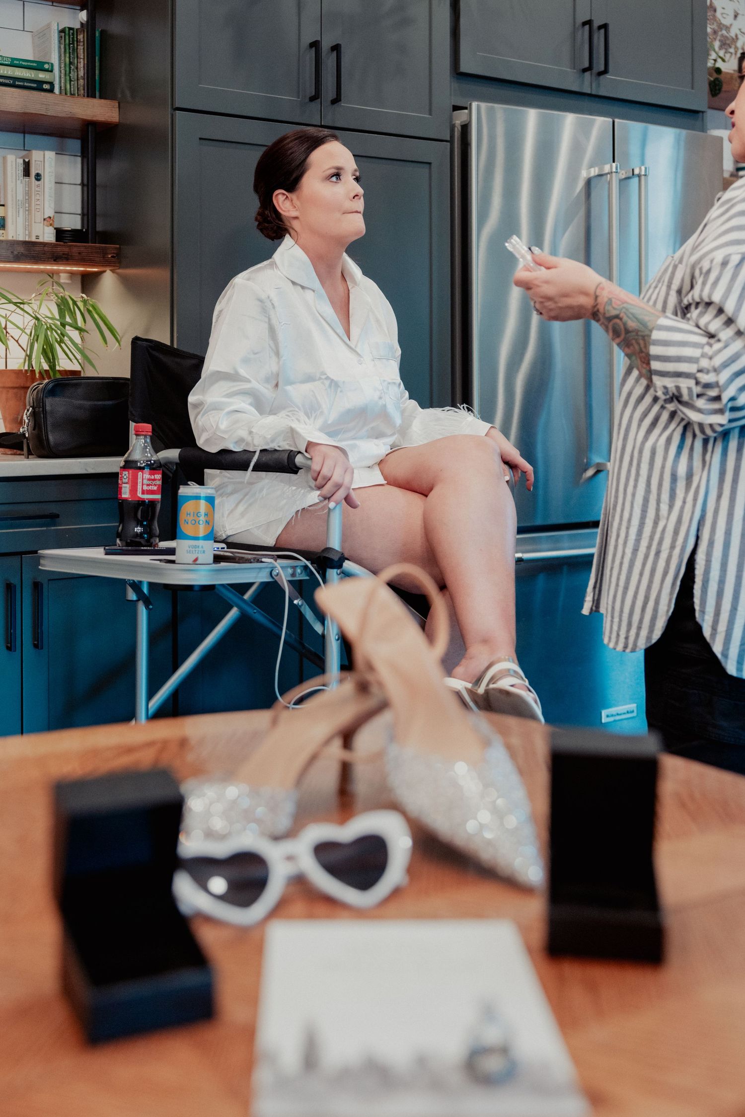 Bride in white robe sitting in kitchen chair with sparkly heels and heart sunglasses in foreground.