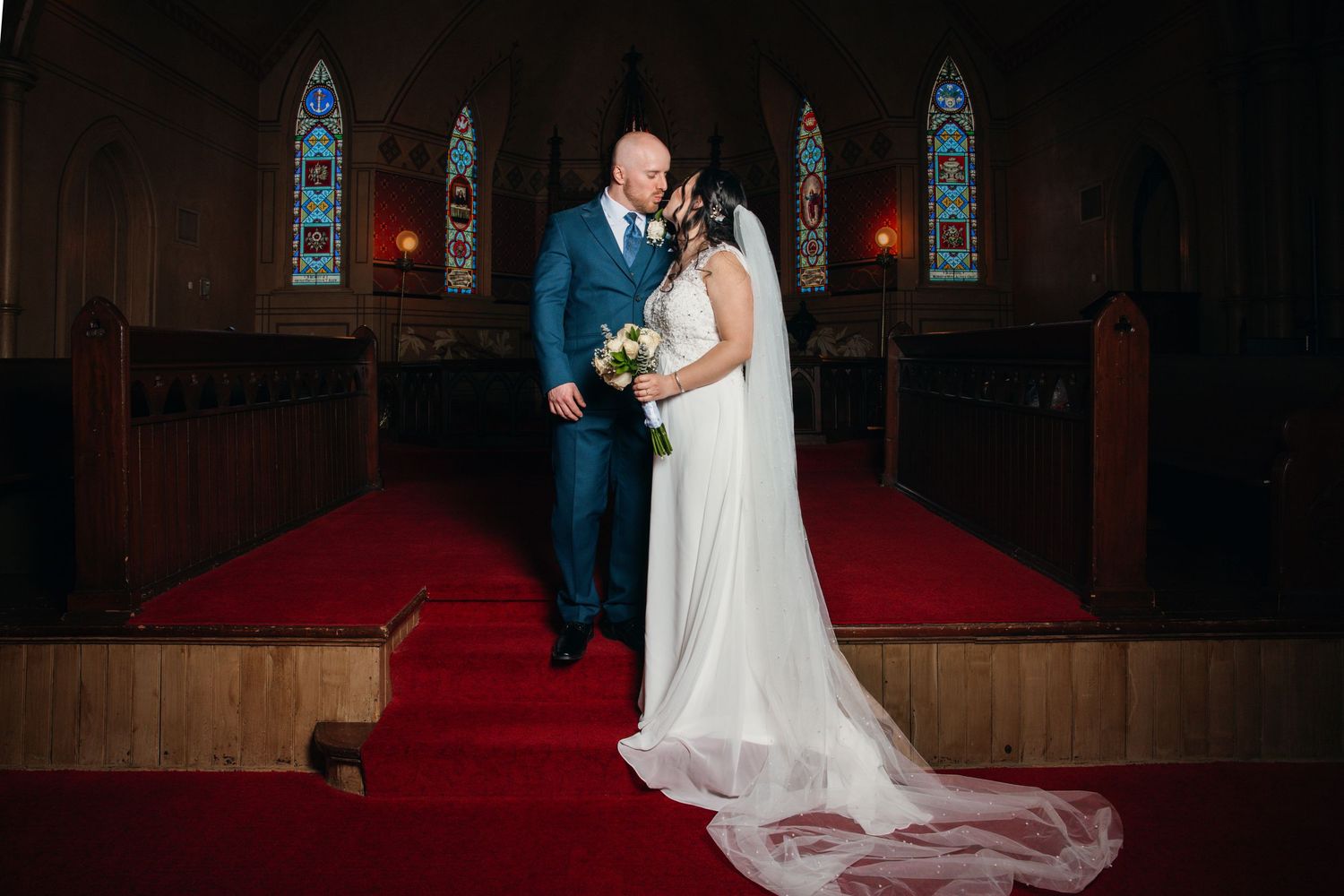 Bride and groom share a tender kiss at the altar of a historic church with red carpet and stained glass windows.
