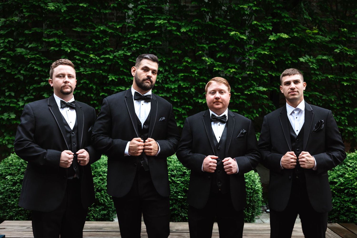 Four men in matching black tuxedos and bow ties posing formally outdoors in front of a green ivy backdrop.