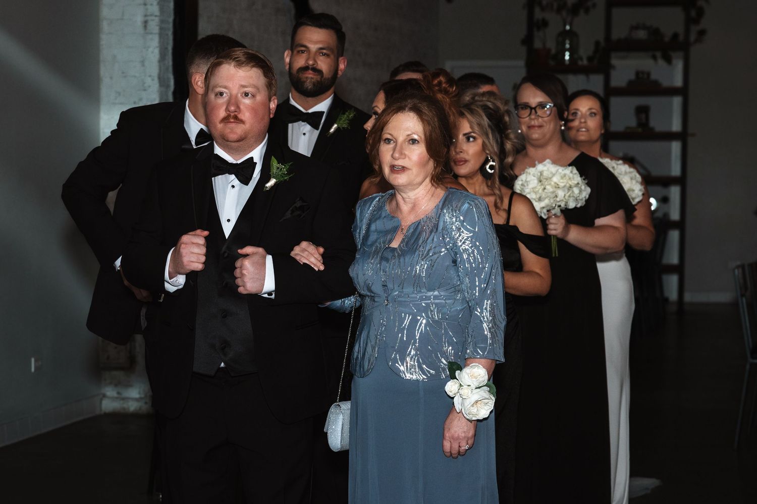 Man in tuxedo escorting elegant woman in blue lace gown during wedding processional.