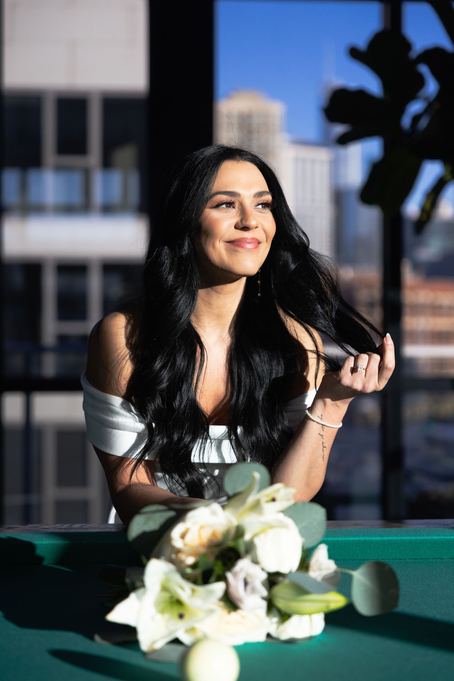 Elegant dark-haired woman in off-shoulder dress leaning on pool table with city view.