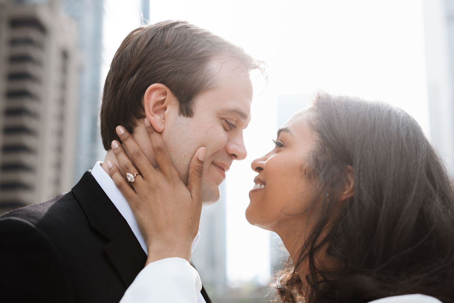 Romantic couple in formal wear sharing a tender moment outdoors with city skyline behind them.