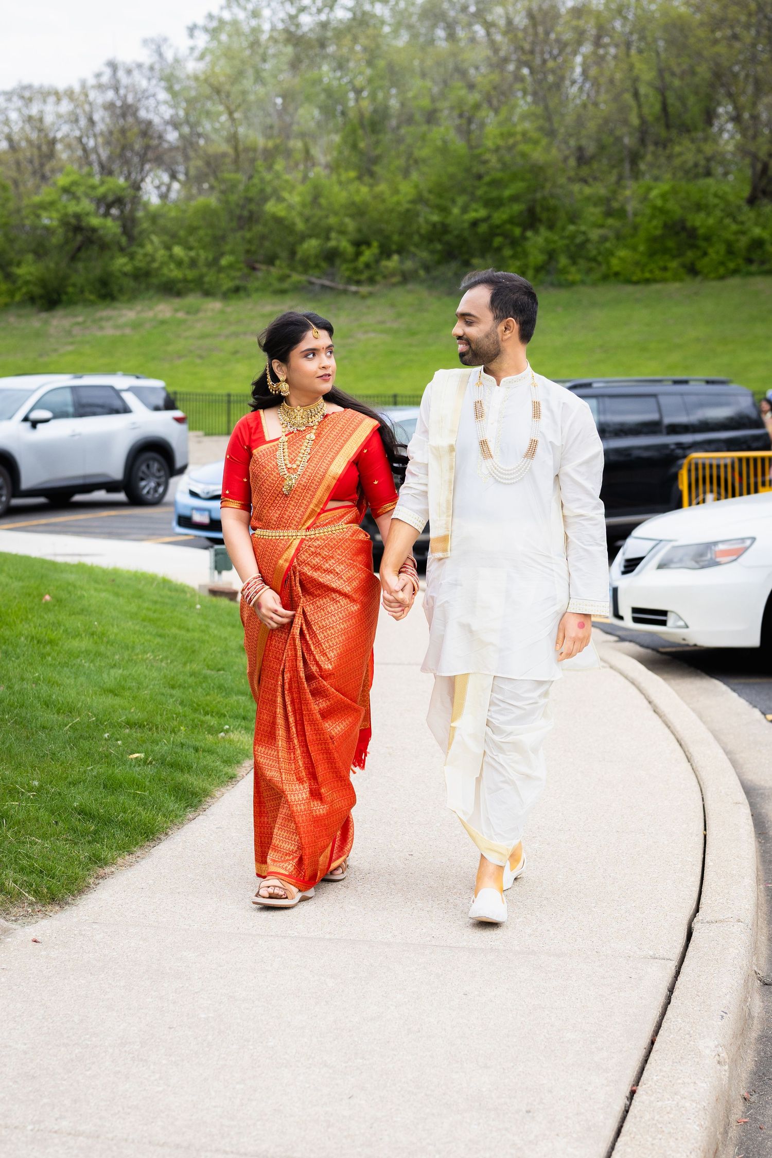 Indian couple in traditional attire walking hand in hand, woman in red saree and man in white kurta.