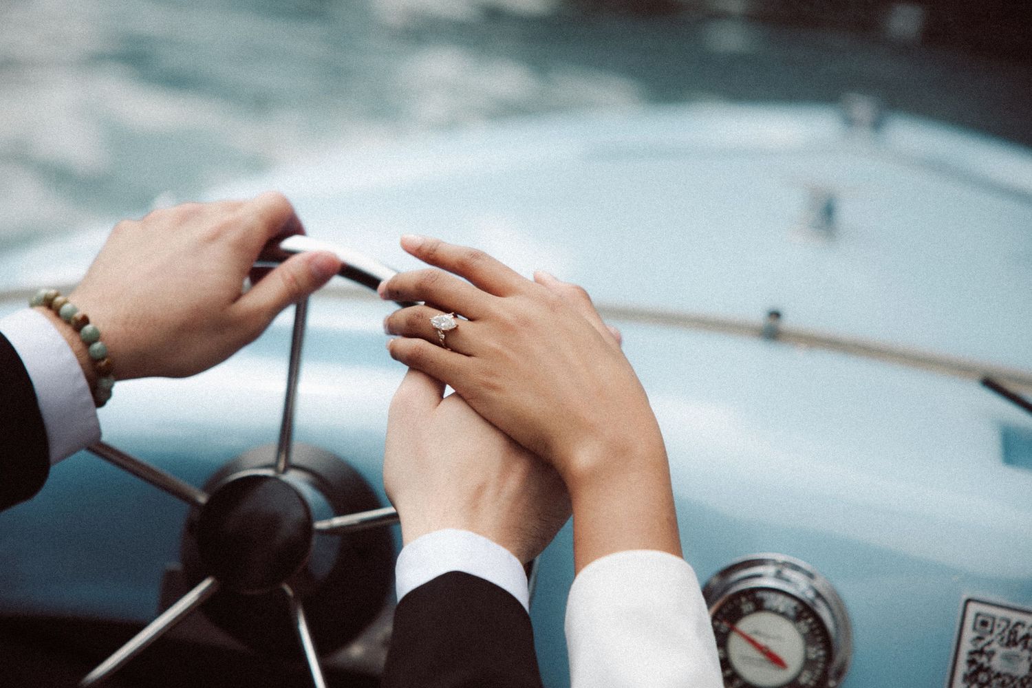 Close-up of couple's hands on a vintage blue boat steering wheel, bride wearing diamond engagement ring.