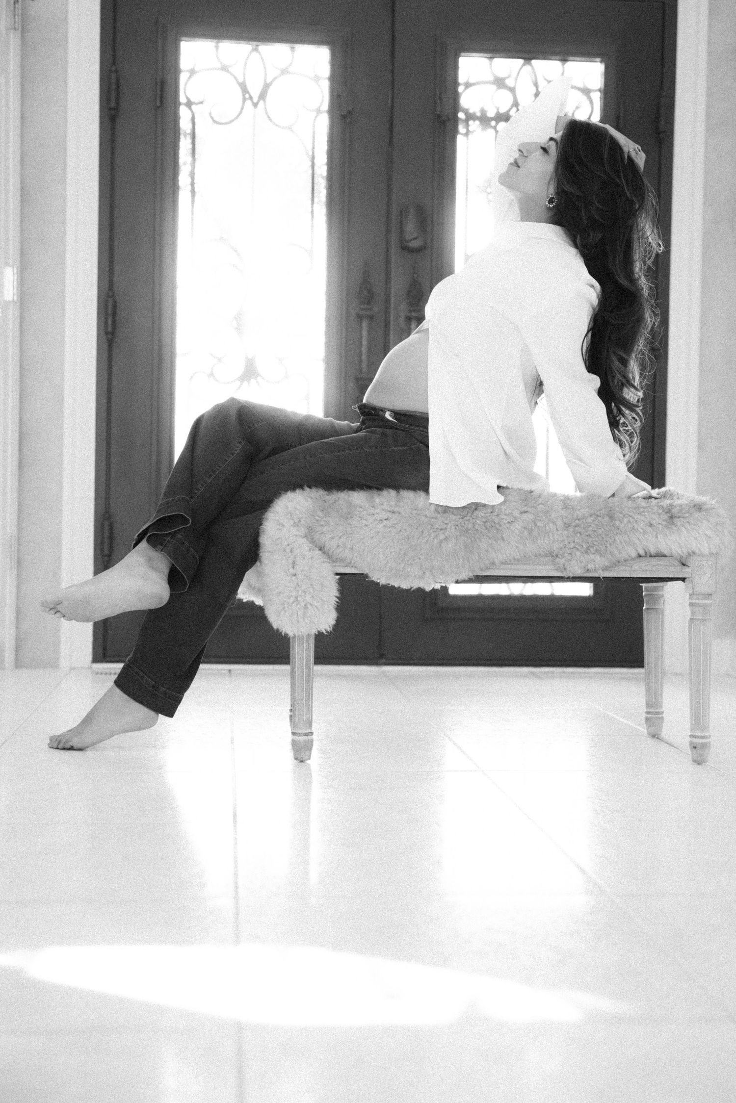 A black and white portrait of a pregnant woman sitting on a bench near elegant front doors in natural light.