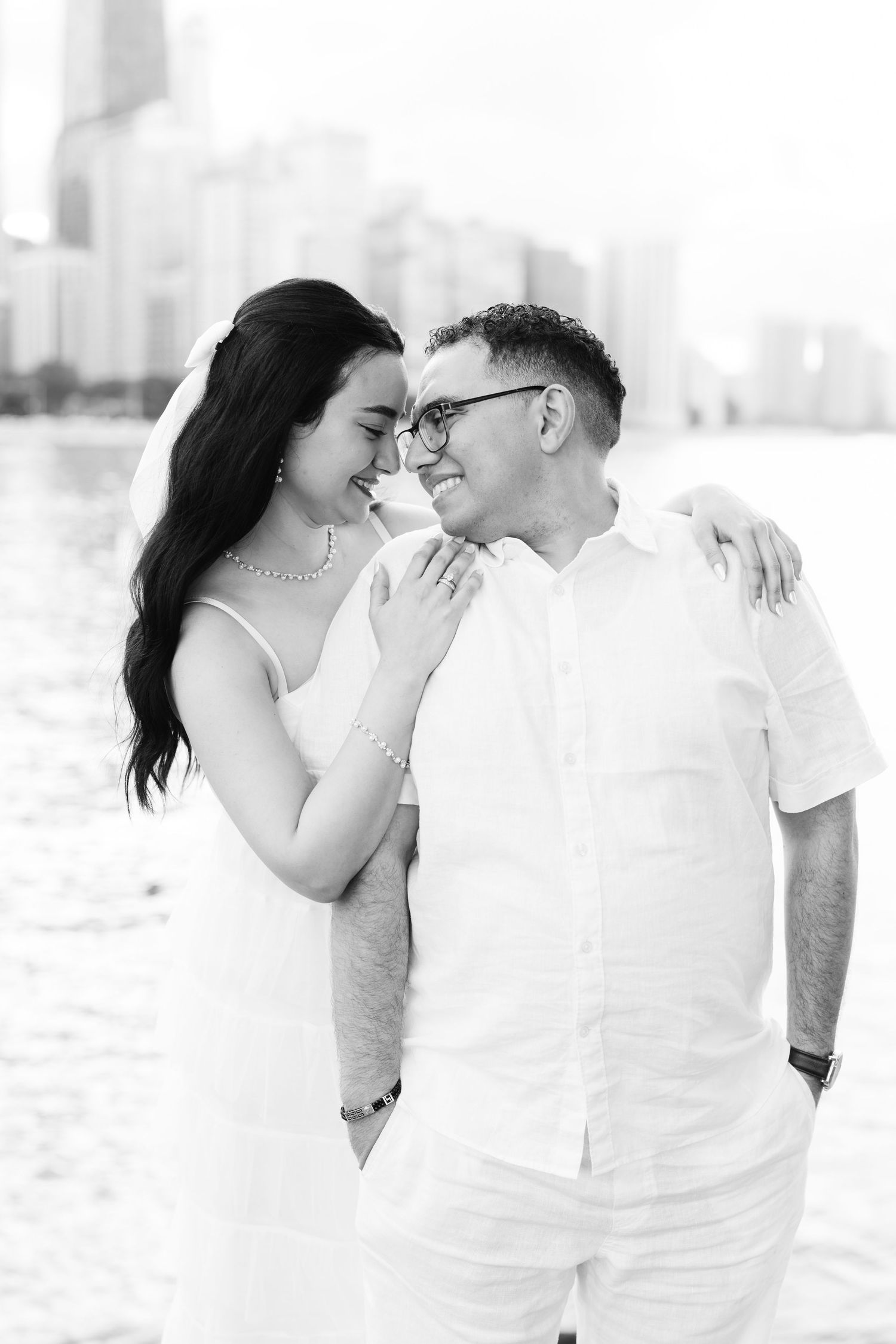 Black and white photo of a smiling couple in white outfits embracing near a waterfront with city skyline behind them.