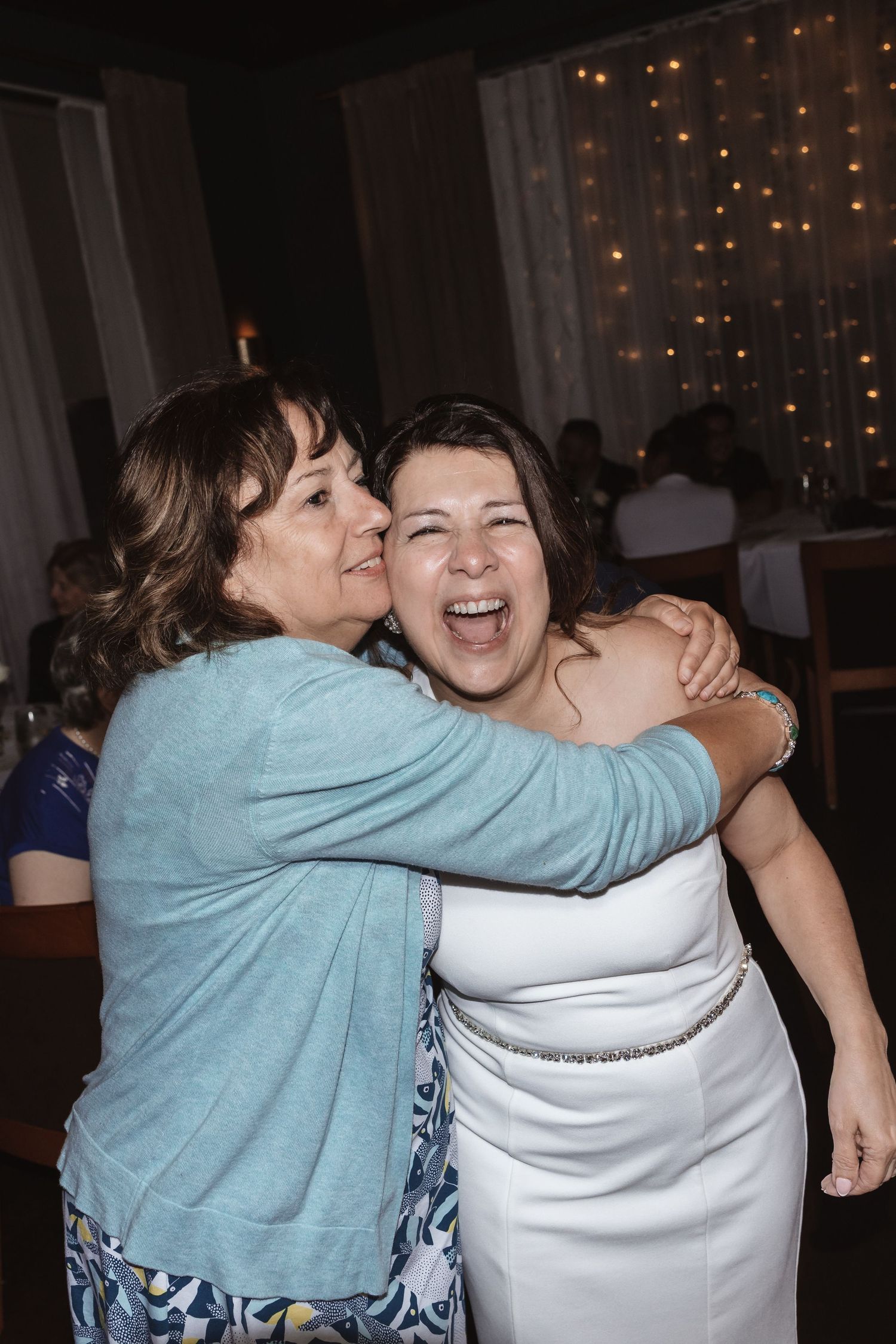Bride in white gown laughs joyfully as a woman in teal hugs her at wedding reception.