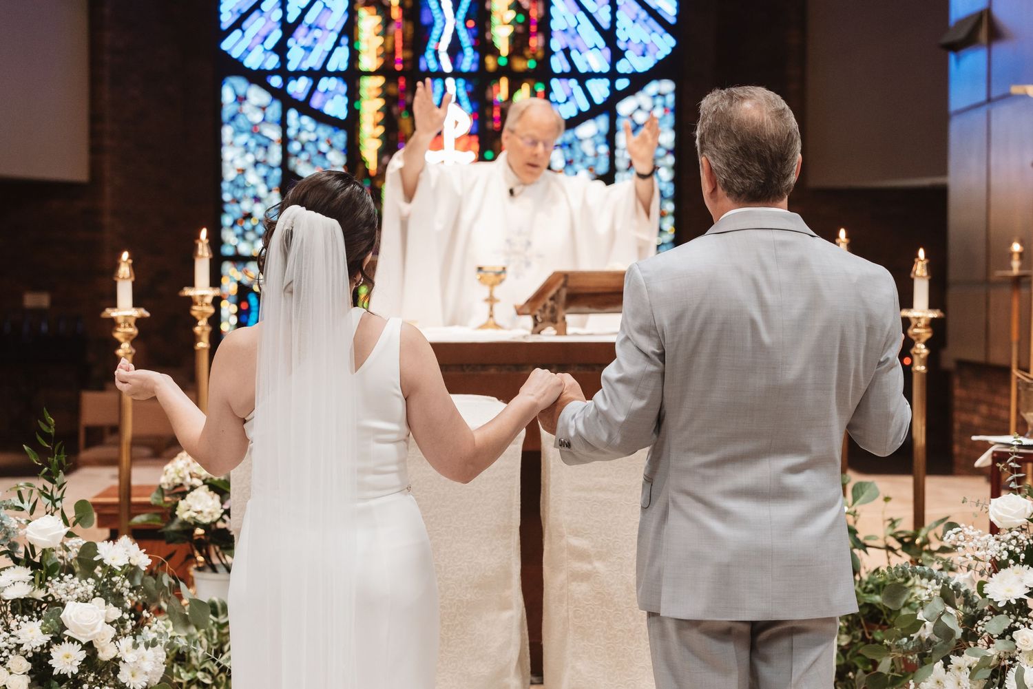 Bride and groom hold hands at church altar before priest during traditional Catholic wedding ceremony with stained glass windows.
