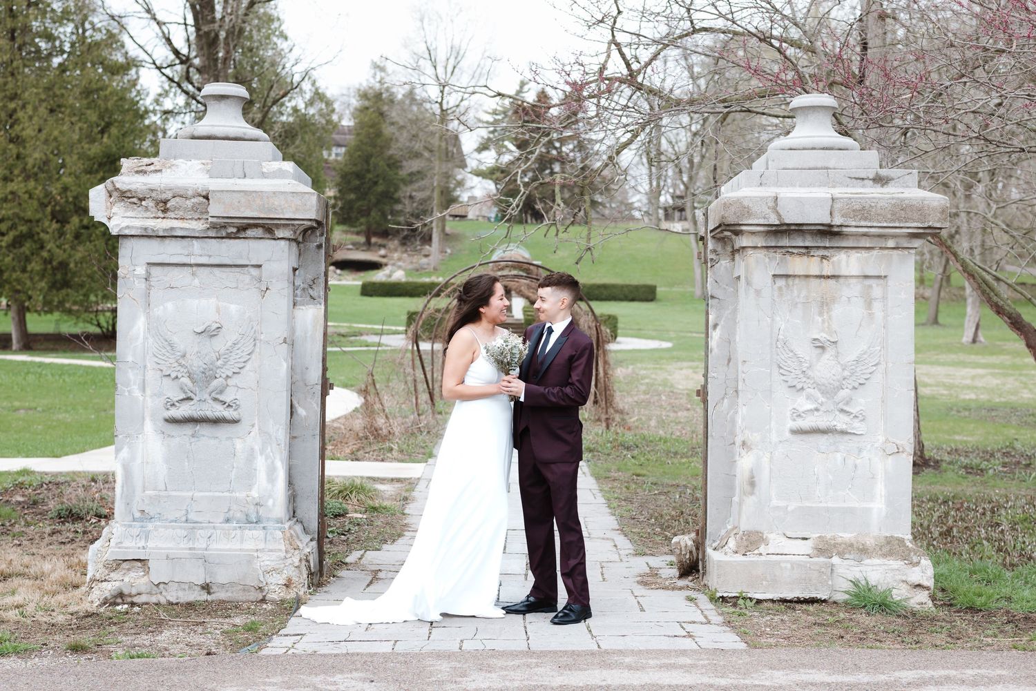 Bride and groom stand between ornate stone gate pillars in a lush green park setting on their wedding day.