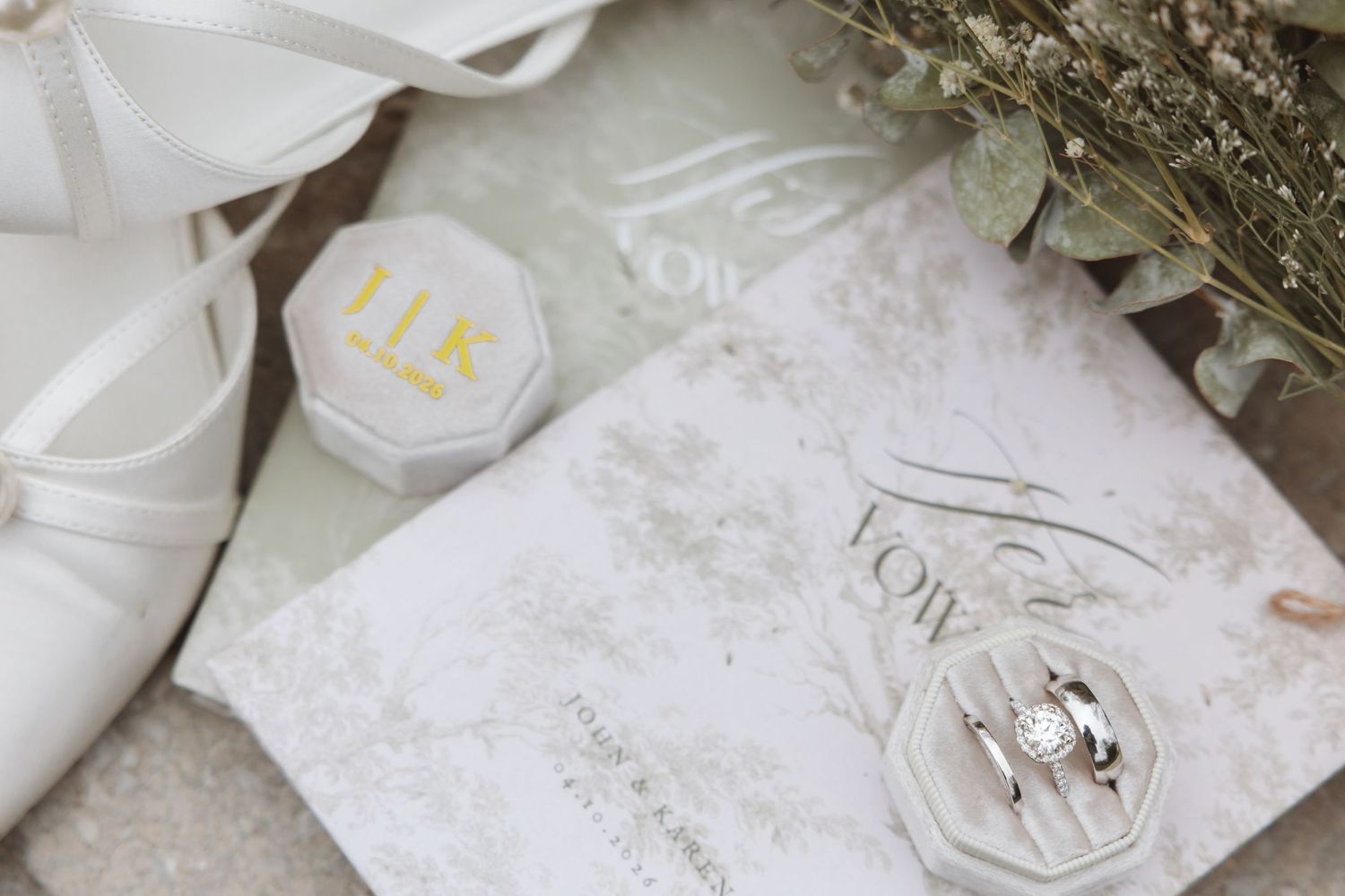 Elegant wedding flat lay featuring bridal shoes, gold monogram ring box, vow books, and diamond rings on a soft green background.