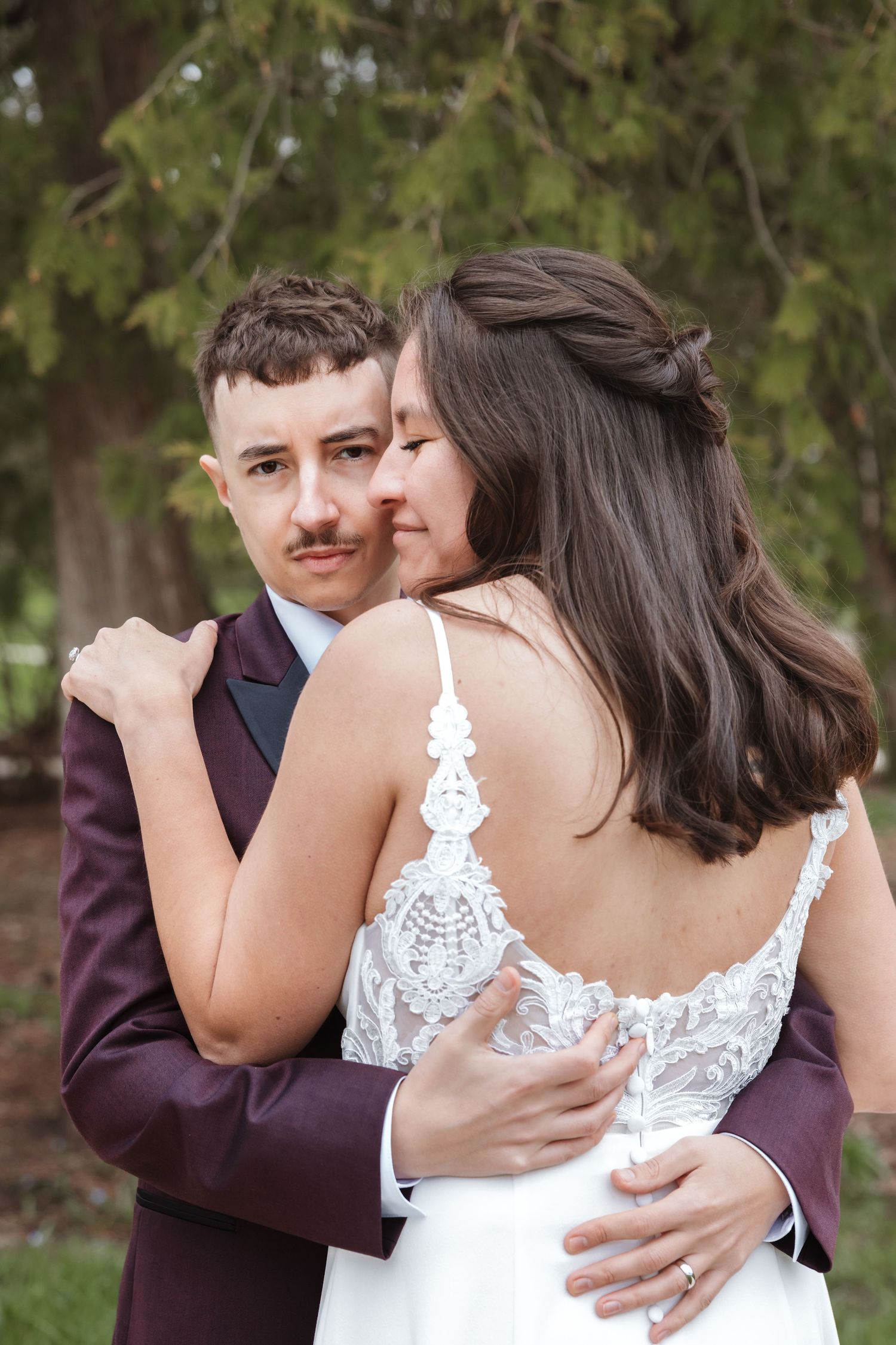 Bride and groom embrace outdoors, she wears lace wedding dress, he wears burgundy suit.