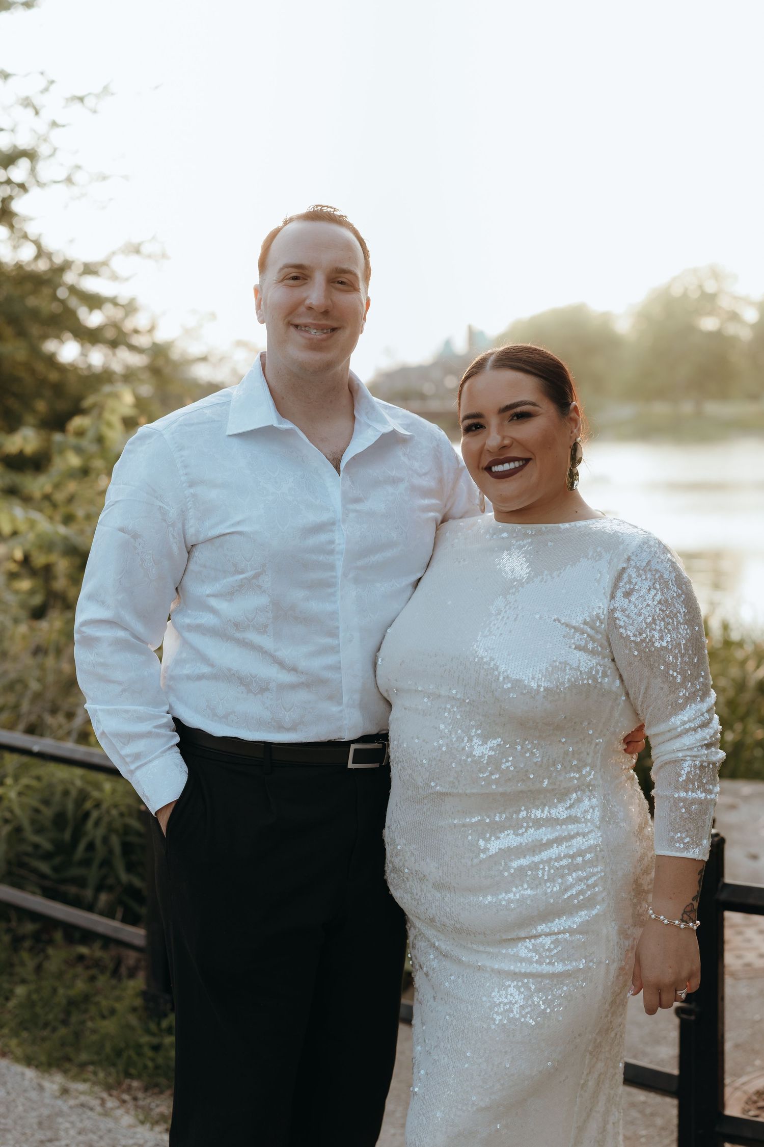 Couple posing outdoors near a lake, bride in sequin gown and groom in white shirt with black pants.