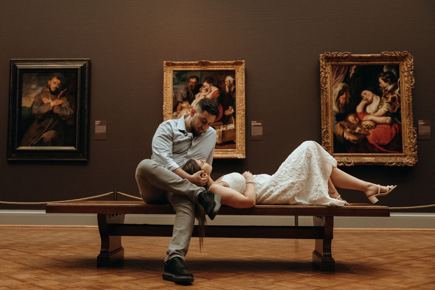 Romantic couple sharing intimate moment on museum bench surrounded by classic paintings in dimly lit gallery.