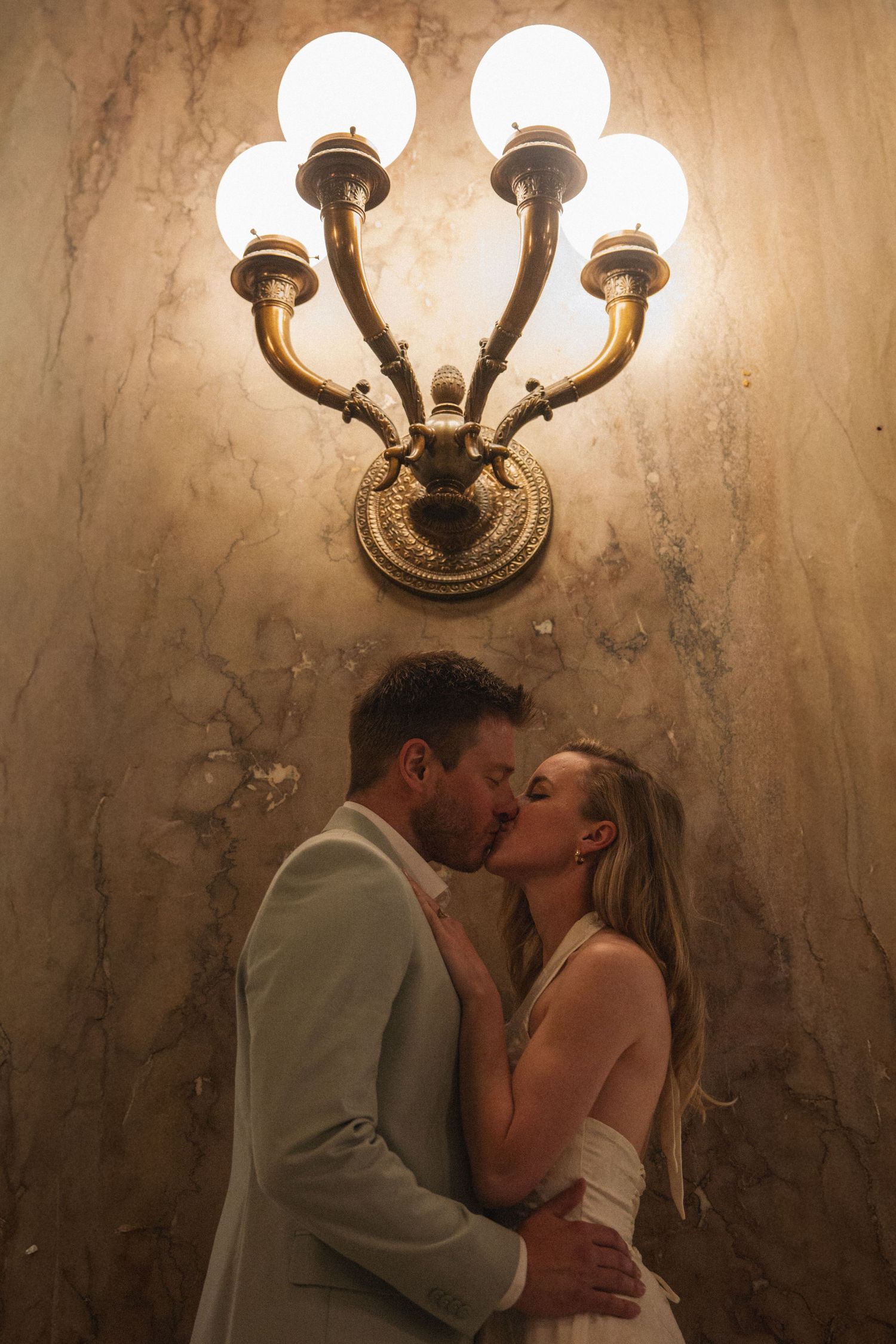 Bride and groom share a passionate kiss beneath a glowing brass wall sconce on a marble wall.