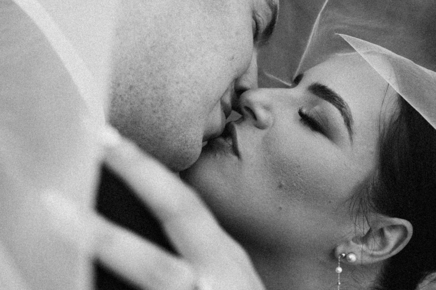 Romantic black and white close-up of newlyweds sharing a tender first kiss beneath a flowing bridal veil.