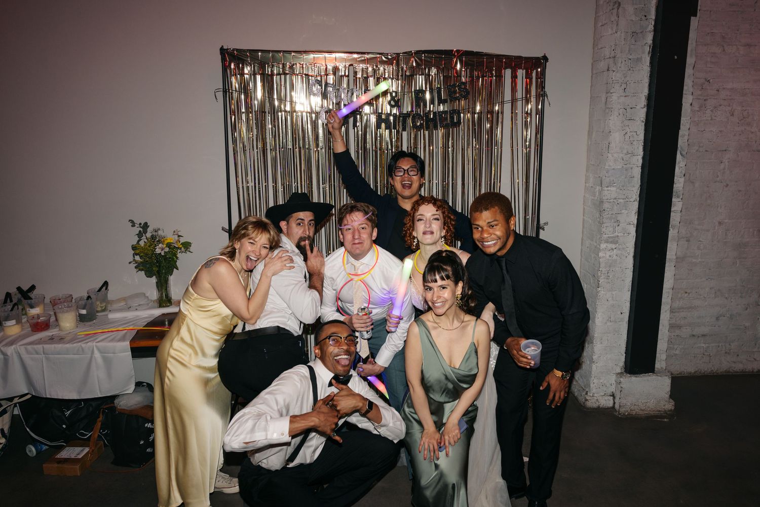 Group of joyful wedding guests posing together in front of a silver tinsel backdrop at a reception.