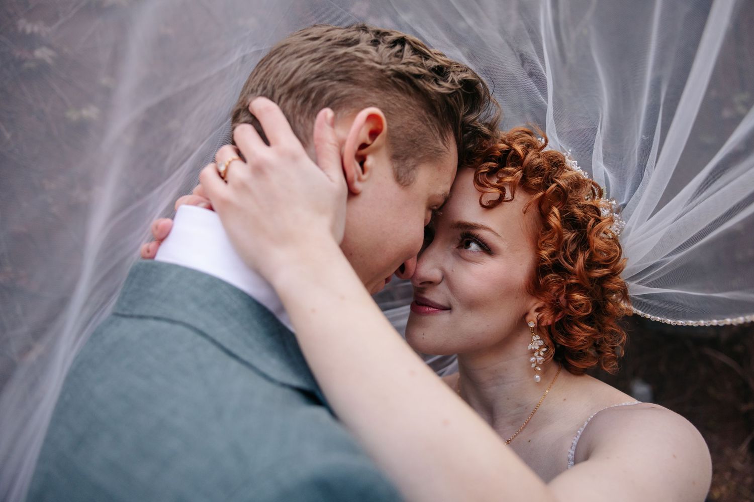 Romantic close-up of bride and groom embracing under wedding veil, bride smiling at camera.