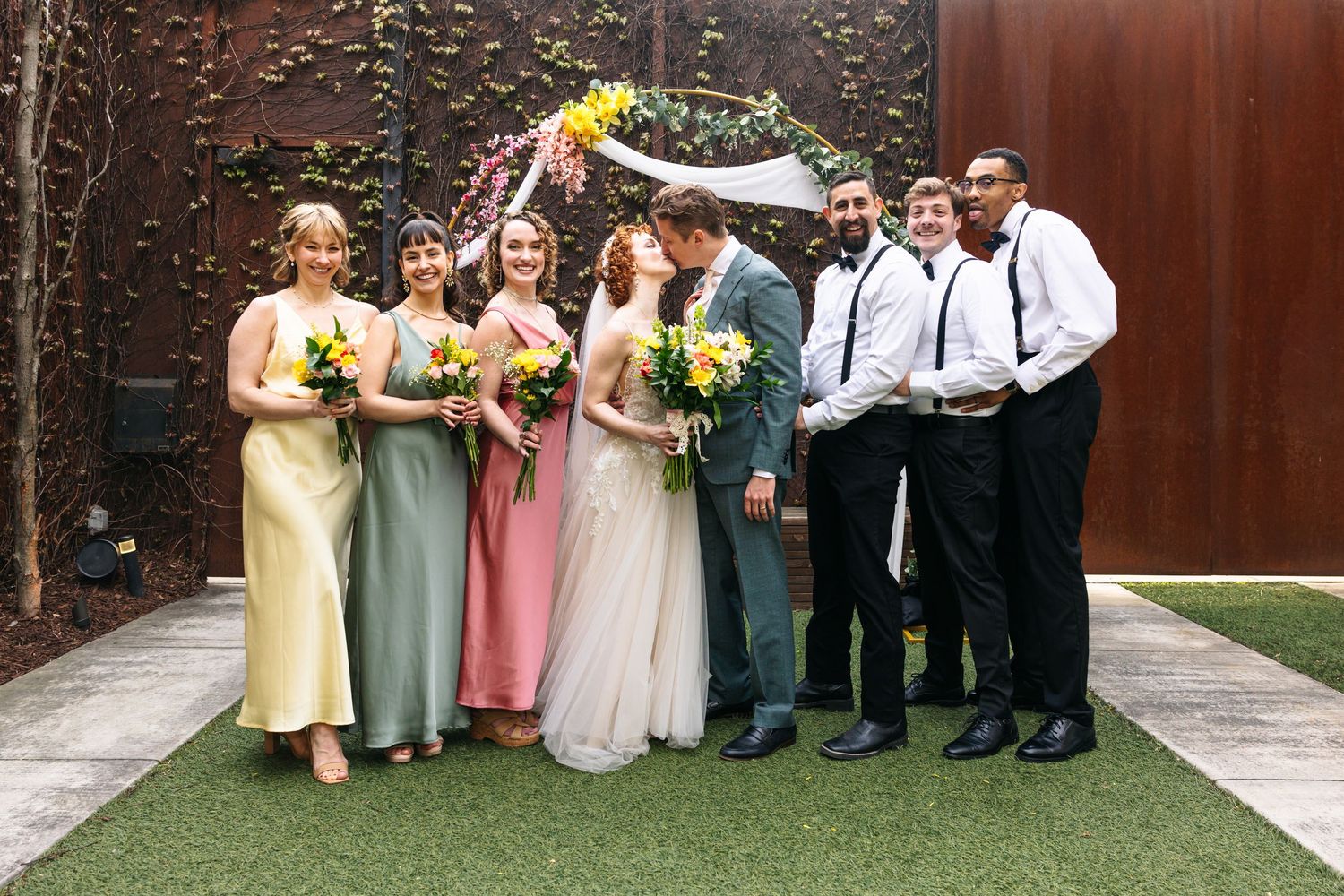 Wedding party poses together outdoors with bride and groom kissing center, bridesmaids in colorful dresses.