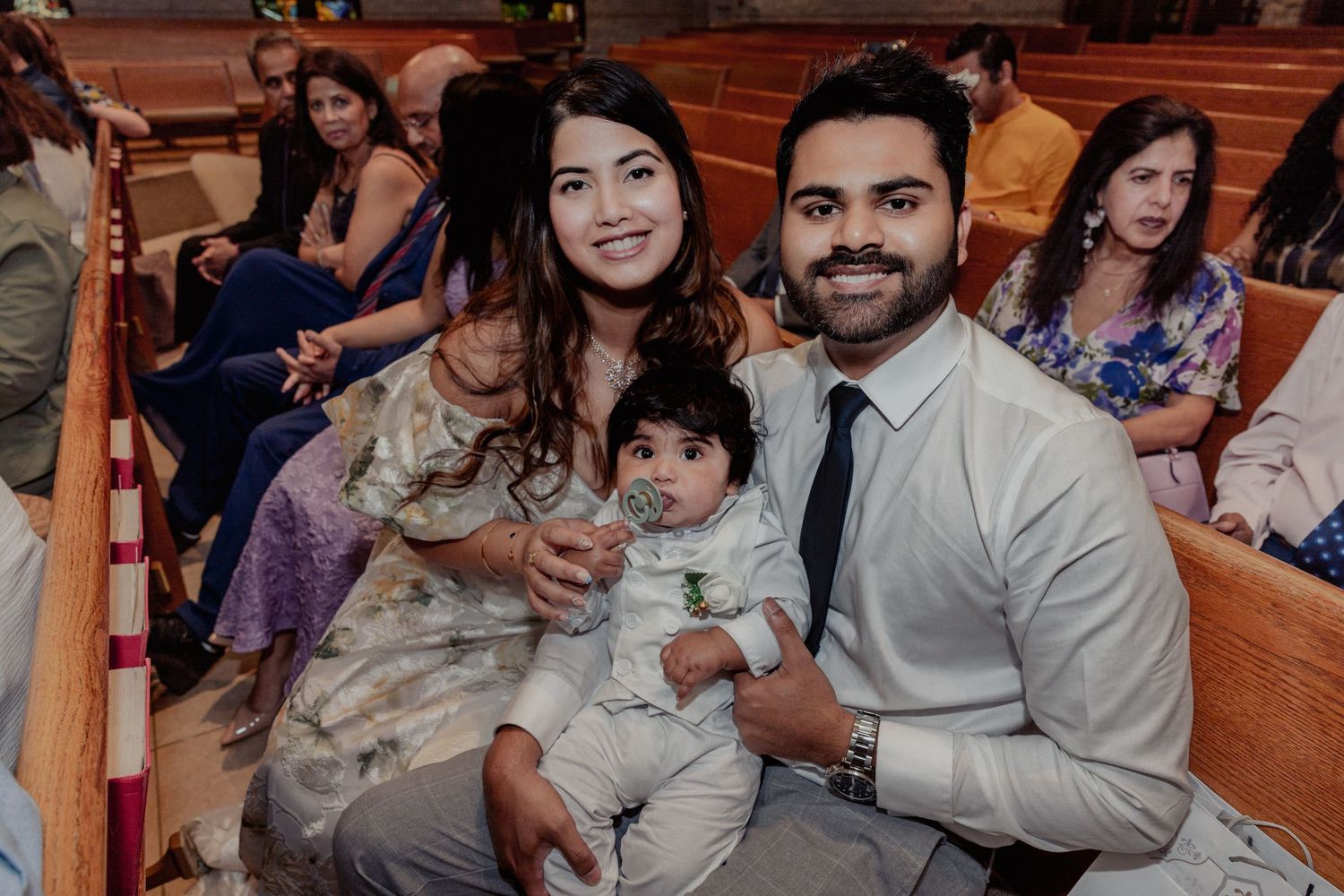 Happy family with baby poses for photo in church pew during baptism celebration.