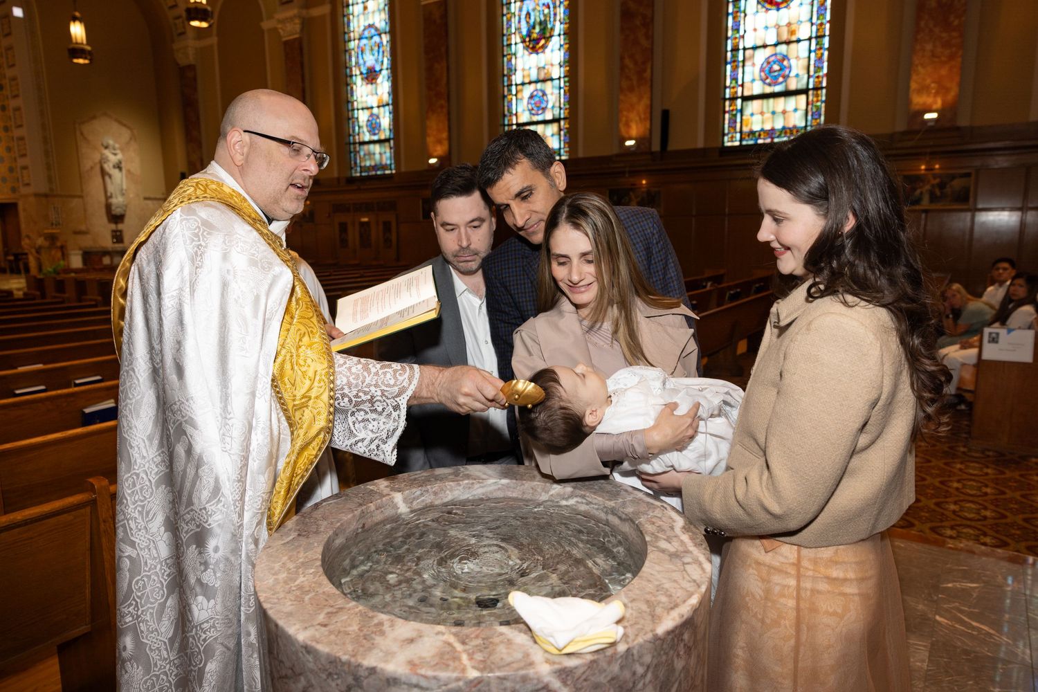 Priest and family gather around a stone baptismal font during an infant's Catholic baptism ceremony.