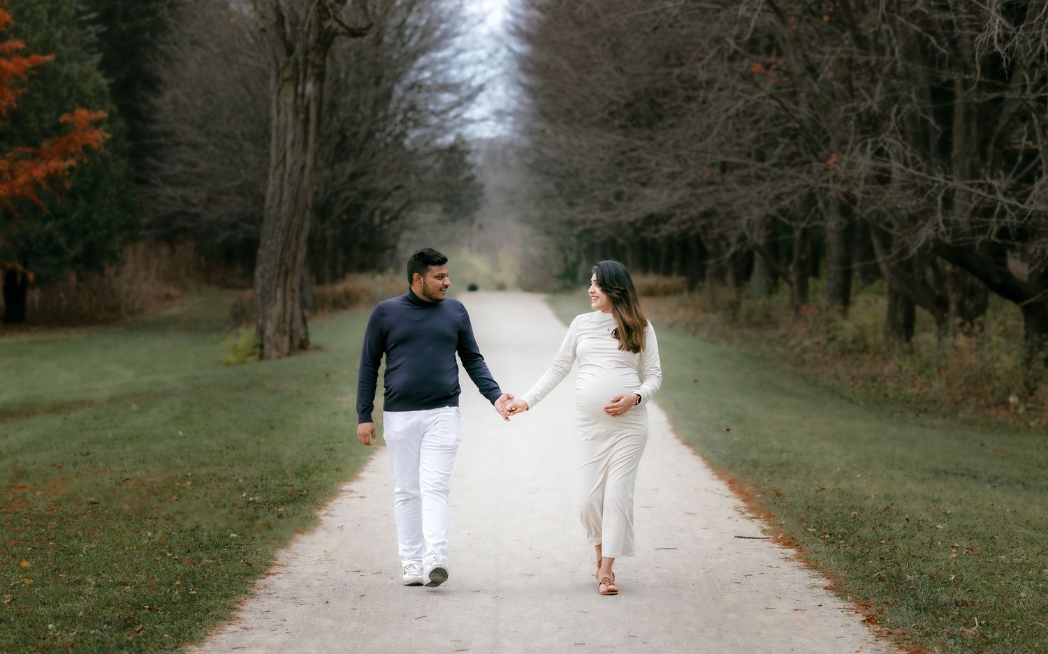 Expecting couple holding hands while walking on tree-lined path during maternity shoot.