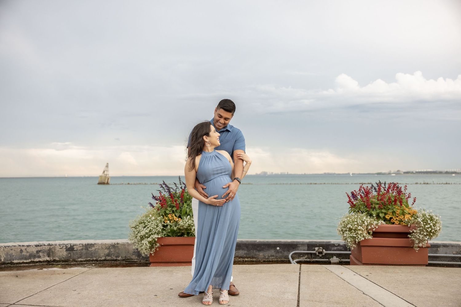 Couple sharing tender moment outdoors, pregnant woman in blue halter maxi dress, man embracing her by the waterfront.
