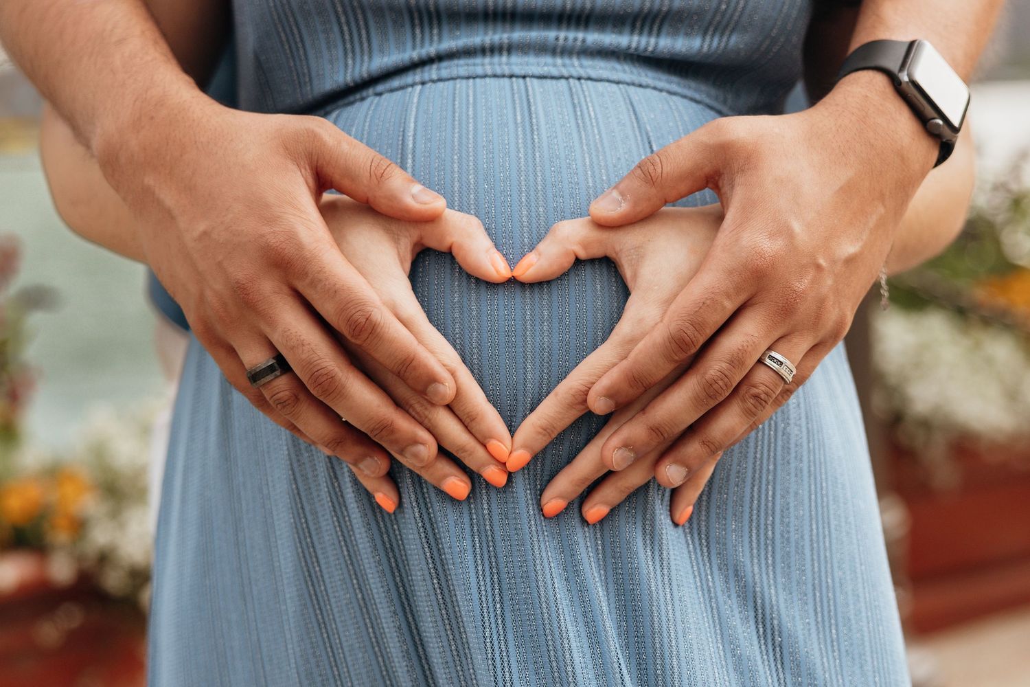 Couple with wedding rings forming heart with hands on pregnant woman's belly in blue maternity dress.