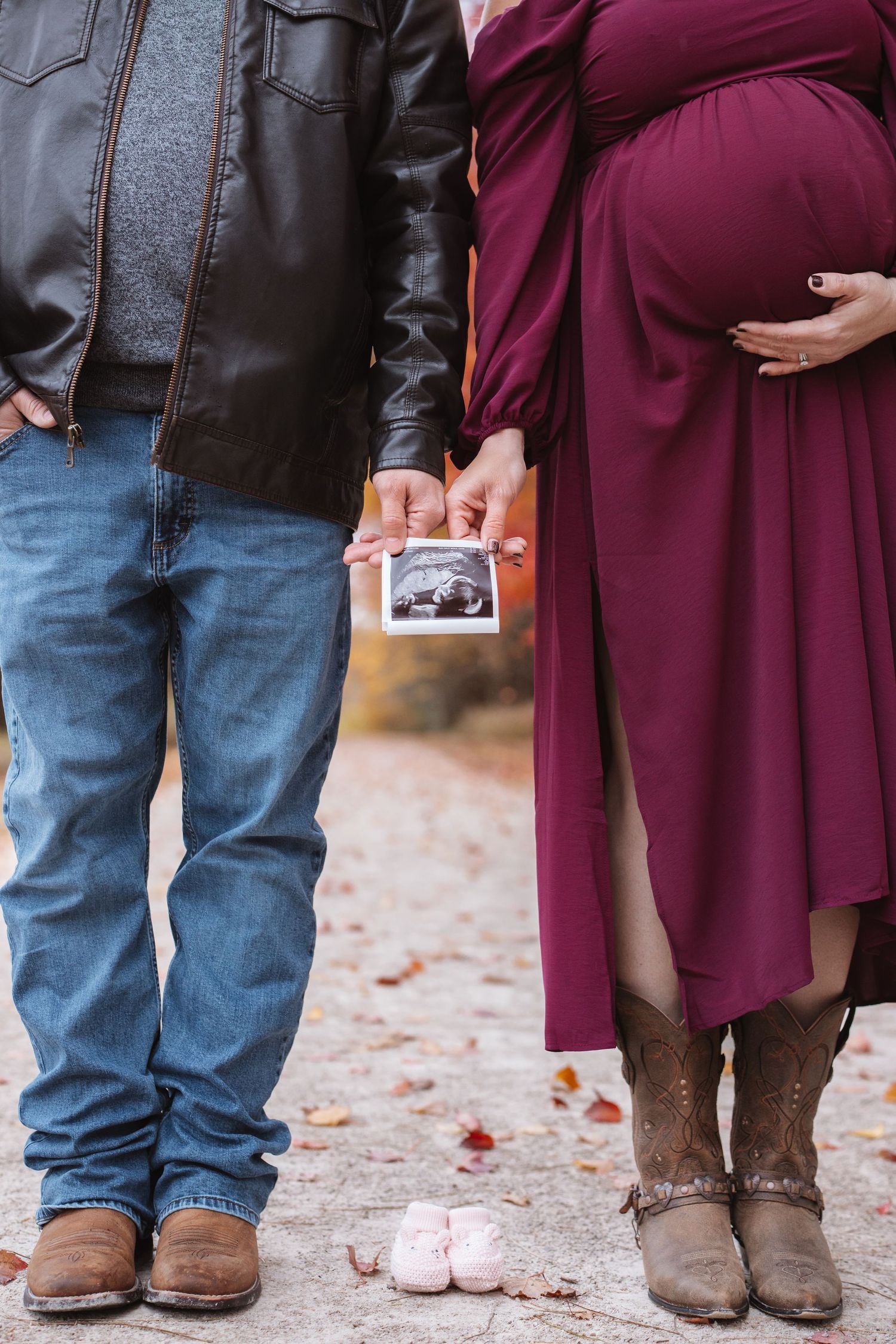 Pregnant couple holding ultrasound photo with tiny pink baby shoes on ground in fall setting.