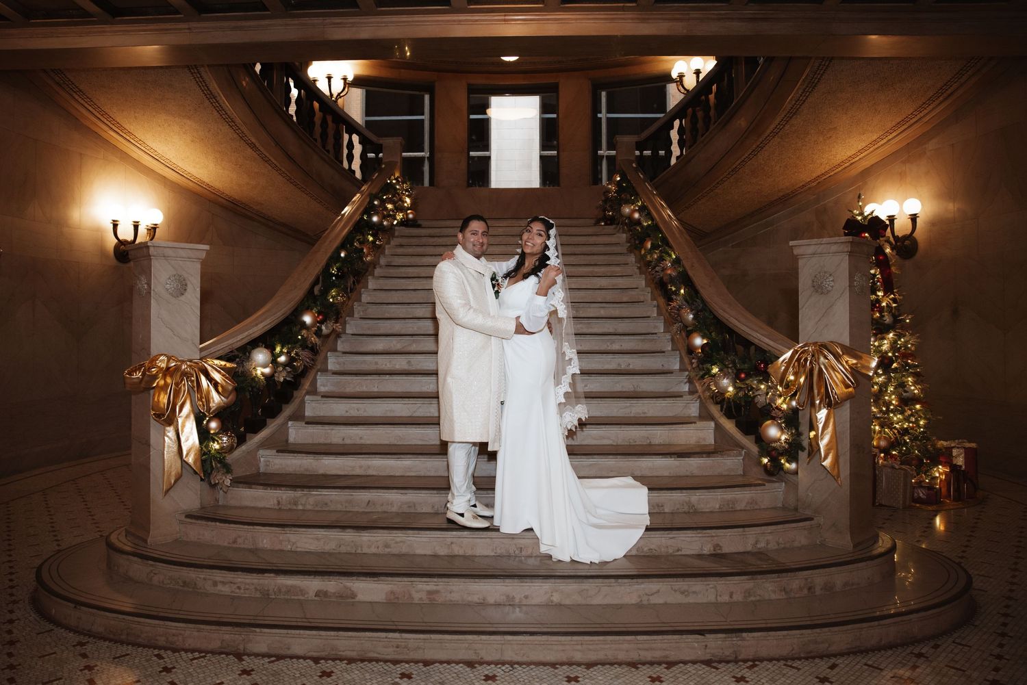 Bride and groom standing on grand marble staircase adorned with festive Christmas garland and gold bows.