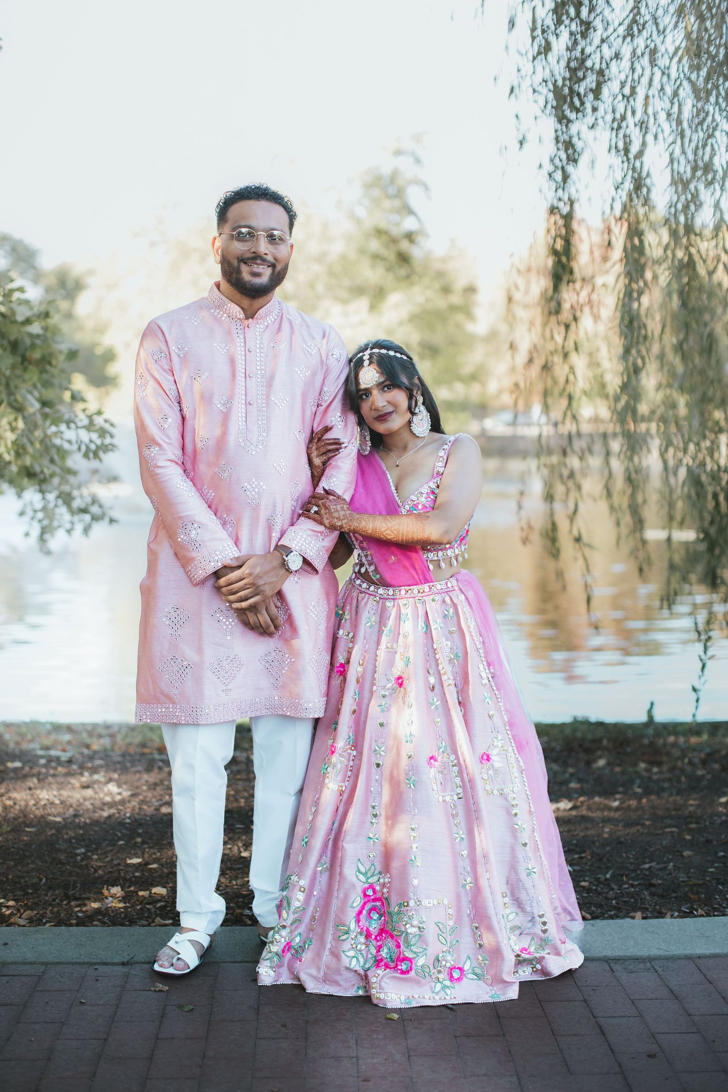 Indian wedding couple posing in pink traditional attire by a serene lake.