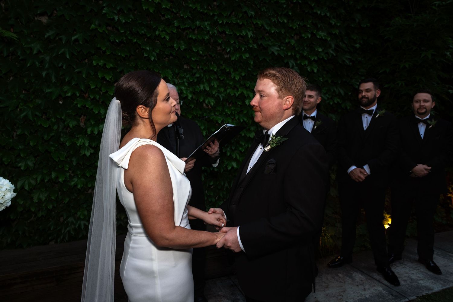 Bride and groom hold hands during wedding vows as officiant reads ceremony before ivy-covered wall.