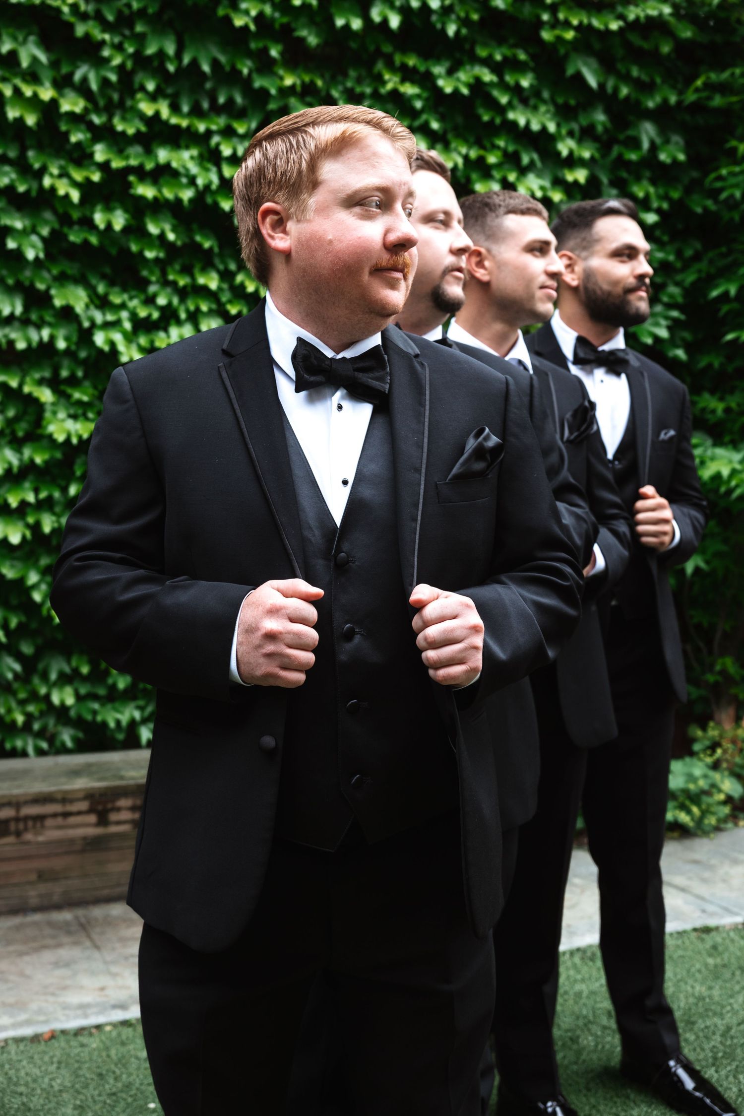 Groom adjusts his tuxedo lapels while groomsmen stand behind him against a green ivy-covered wall.