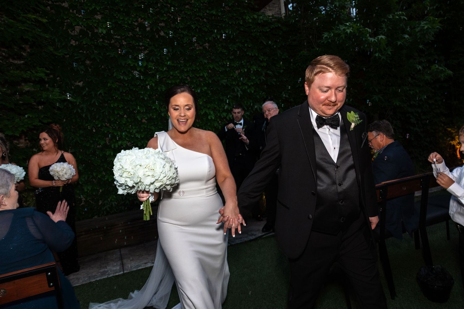 Bride and groom exit wedding ceremony holding hands, guests applauding along the aisle with lush green backdrop.