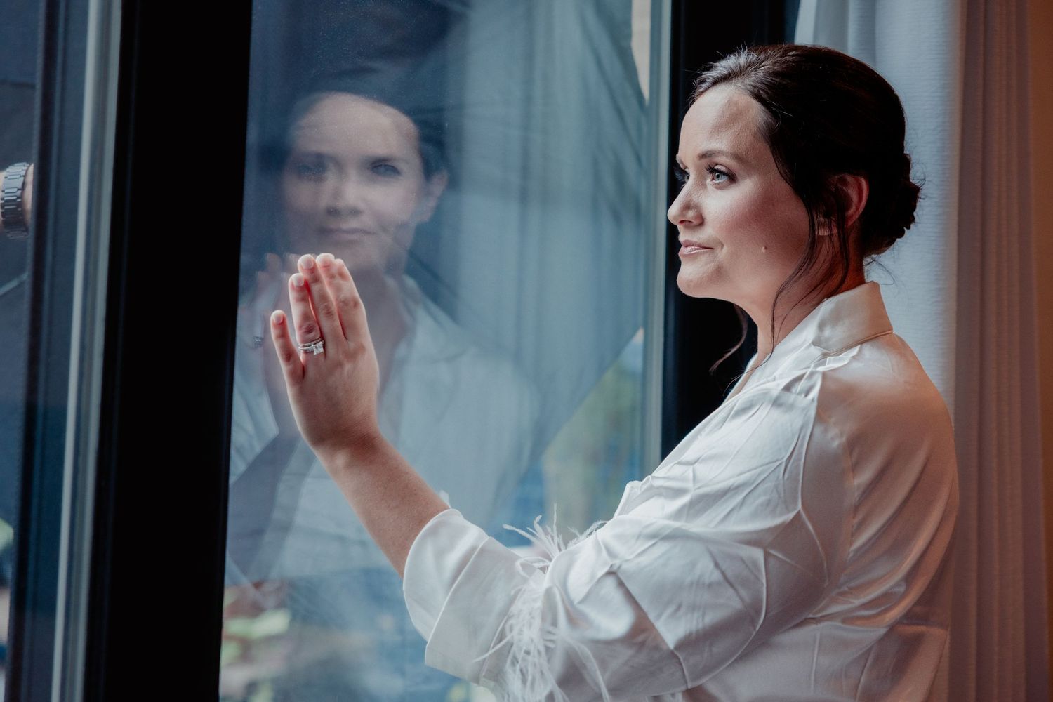 Elegant bride in white robe looking out window, her reflection mirrored in the glass.