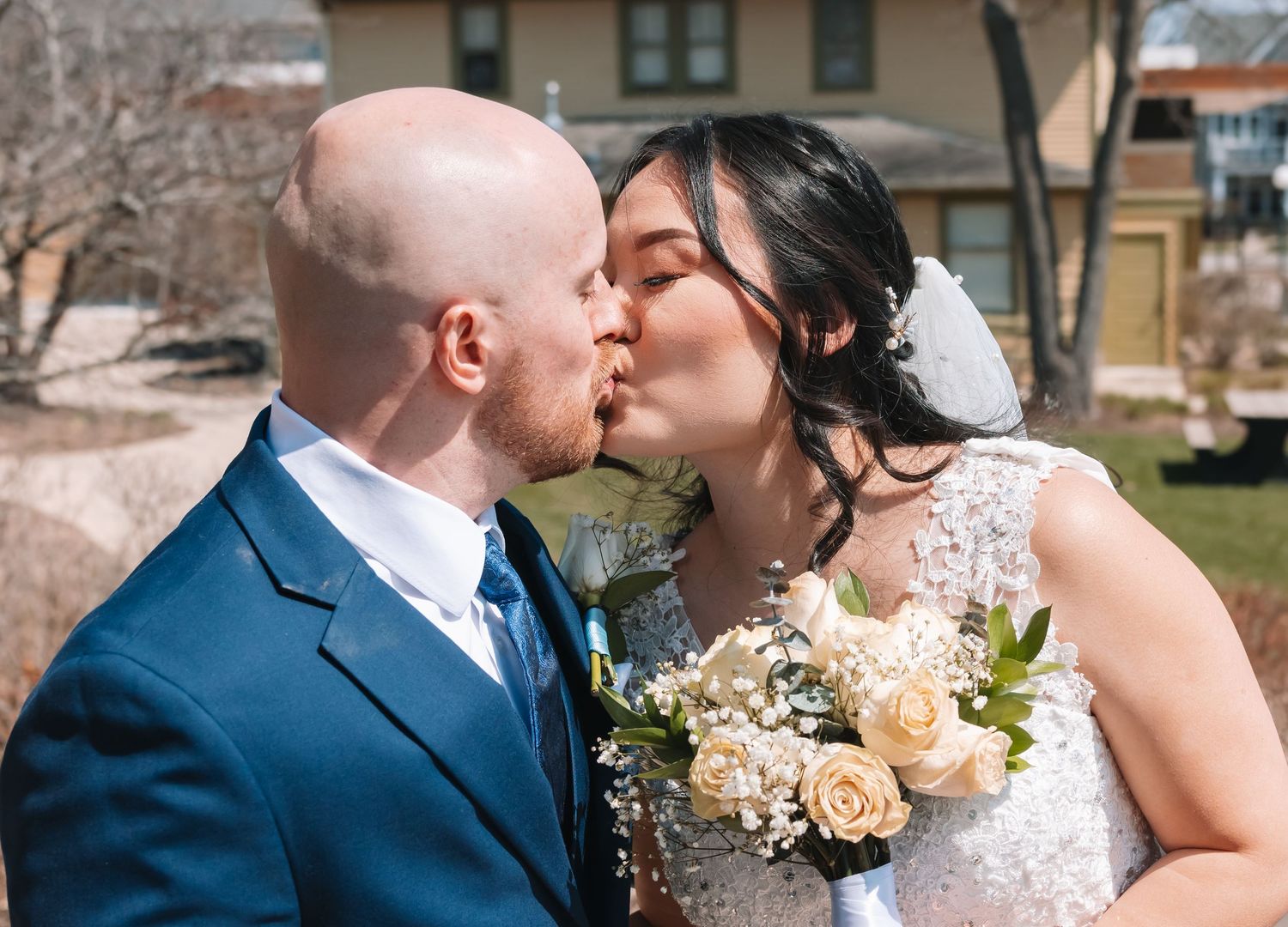 Newlyweds share a joyful kiss outdoors, bride holding a bouquet of cream roses on their wedding day.
