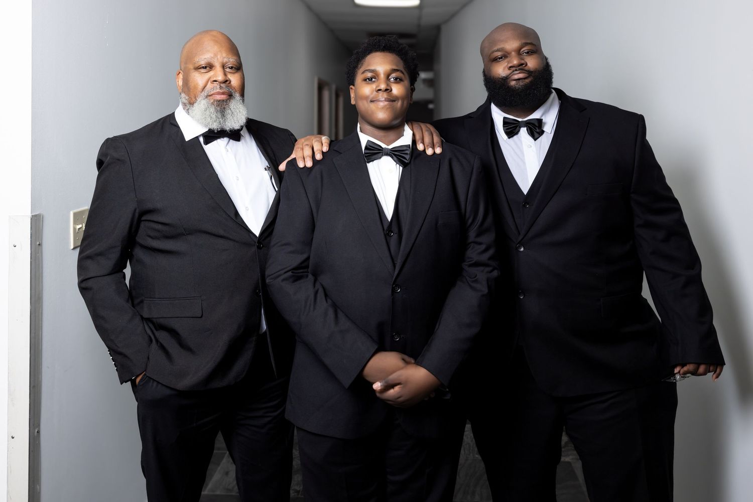 Three men in black tuxedos with bow ties posing together in a hallway for a wedding photo.