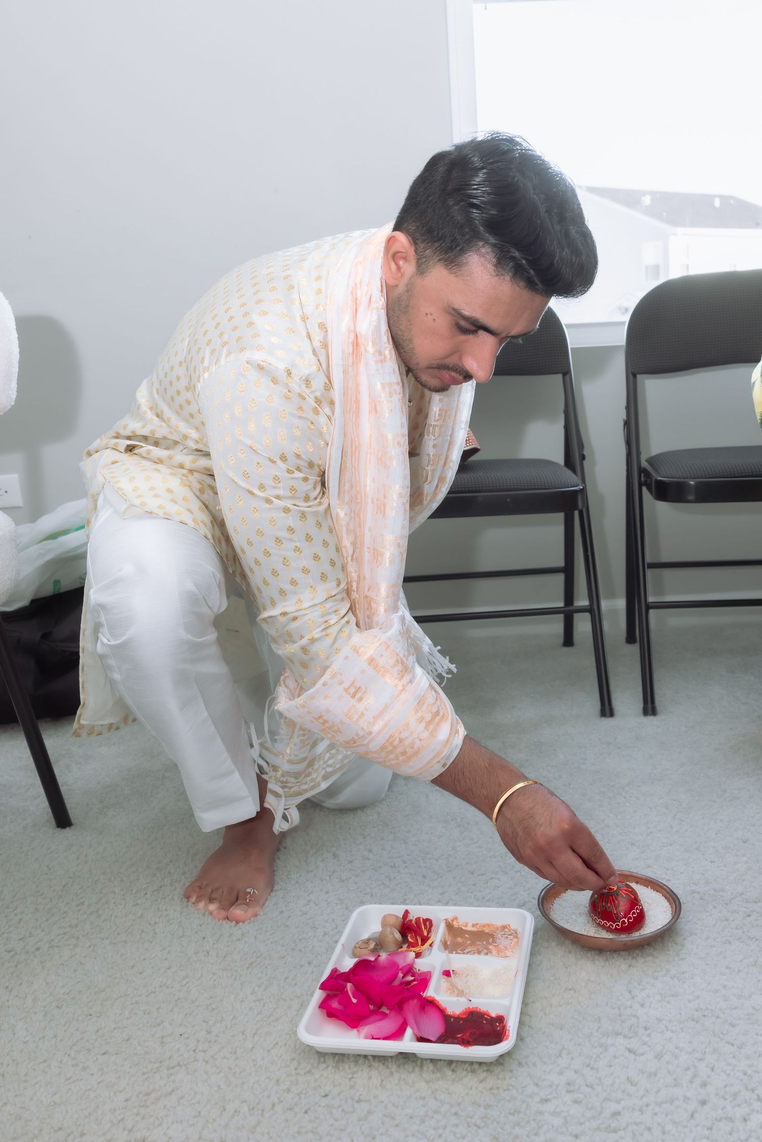 Man in traditional Indian kurta kneeling on floor arranging ritual items on tray during wedding ceremony.