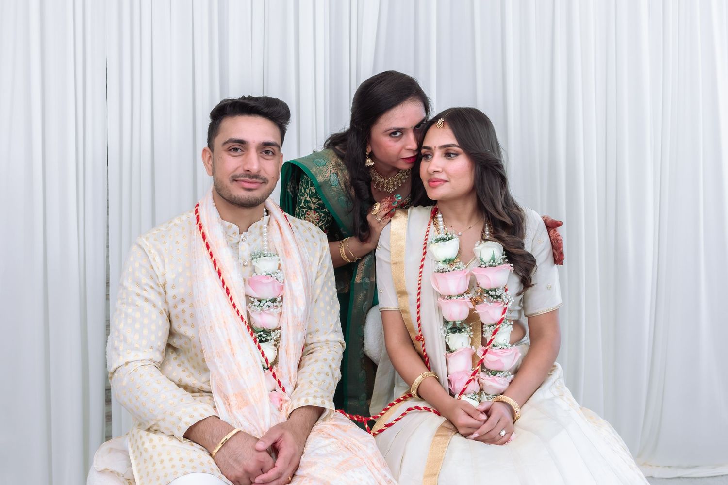Indian bride and groom in garlands share tender moment with woman in green saree.