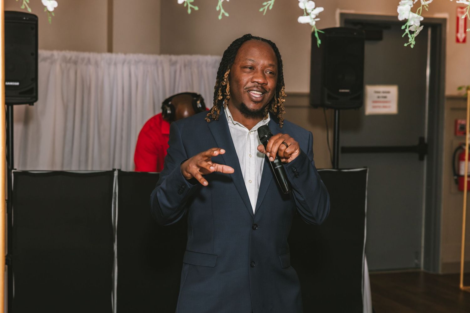 Man in navy suit speaking into microphone at wedding reception with speakers in background.