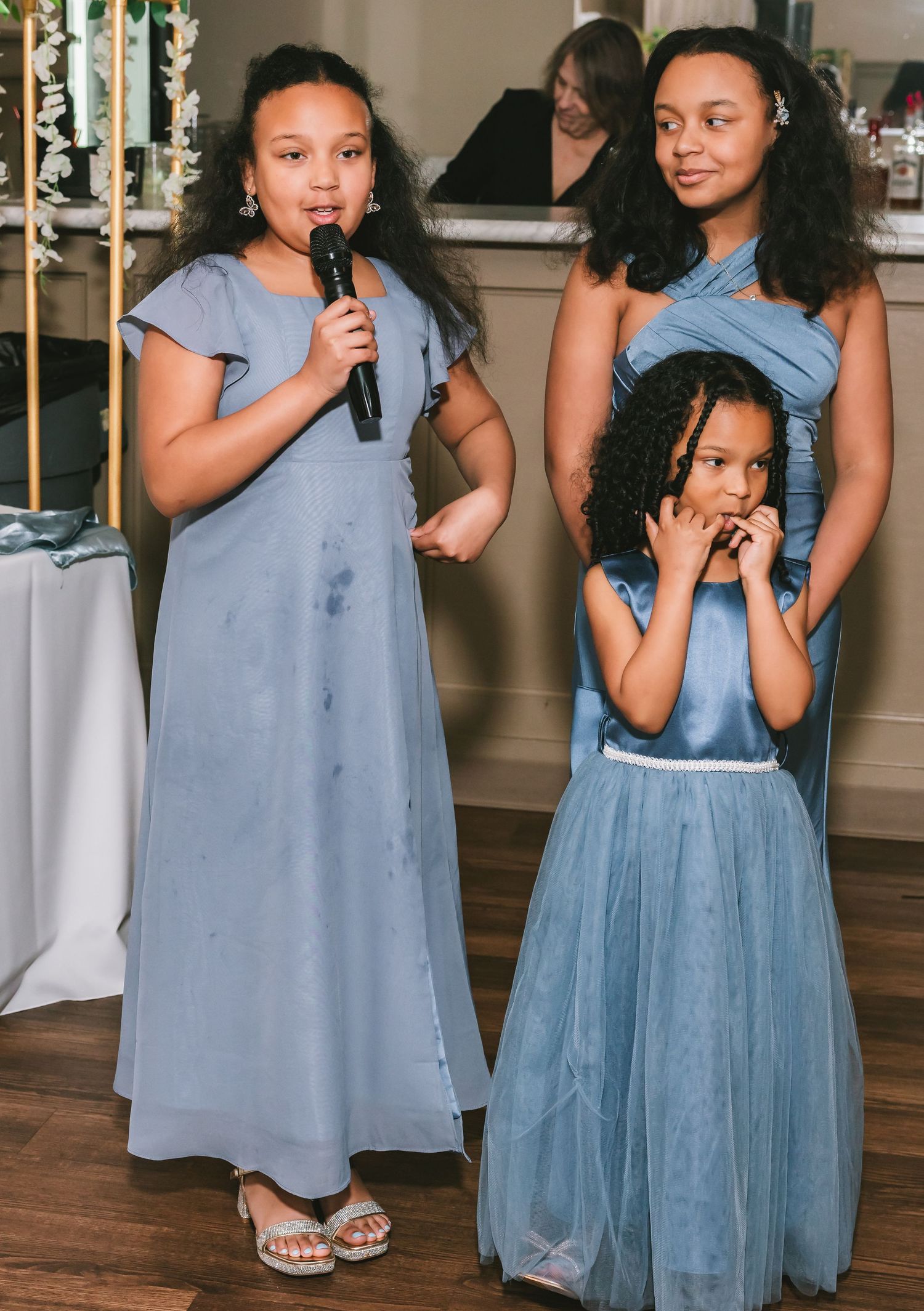 Three girls in dusty blue dresses, one speaking into microphone, at a wedding reception.