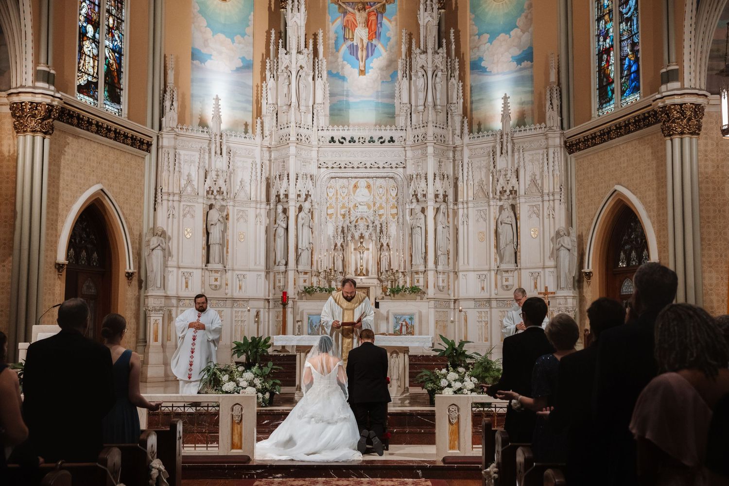 Wedding ceremony at ornate Catholic church altar with bride in white gown and groom before priest.