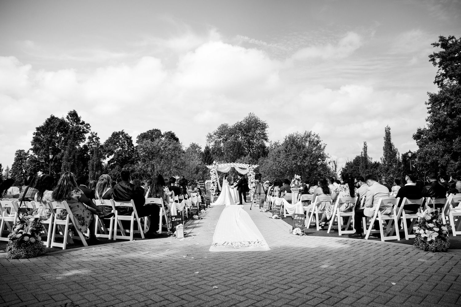Black and white wide shot of outdoor wedding ceremony with white chairs and floral arch.
