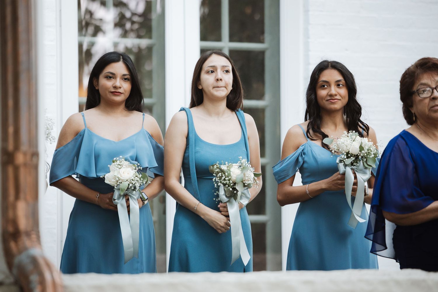 Three bridesmaids in matching teal cold-shoulder dresses holding white ribbon bouquets at outdoor wedding.