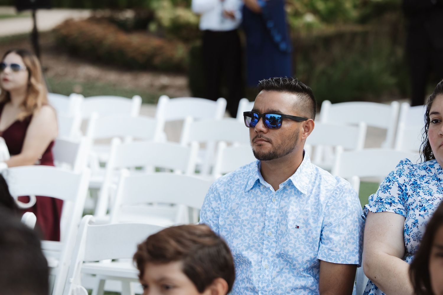 Man in blue floral shirt and sunglasses seated at an outdoor wedding ceremony.