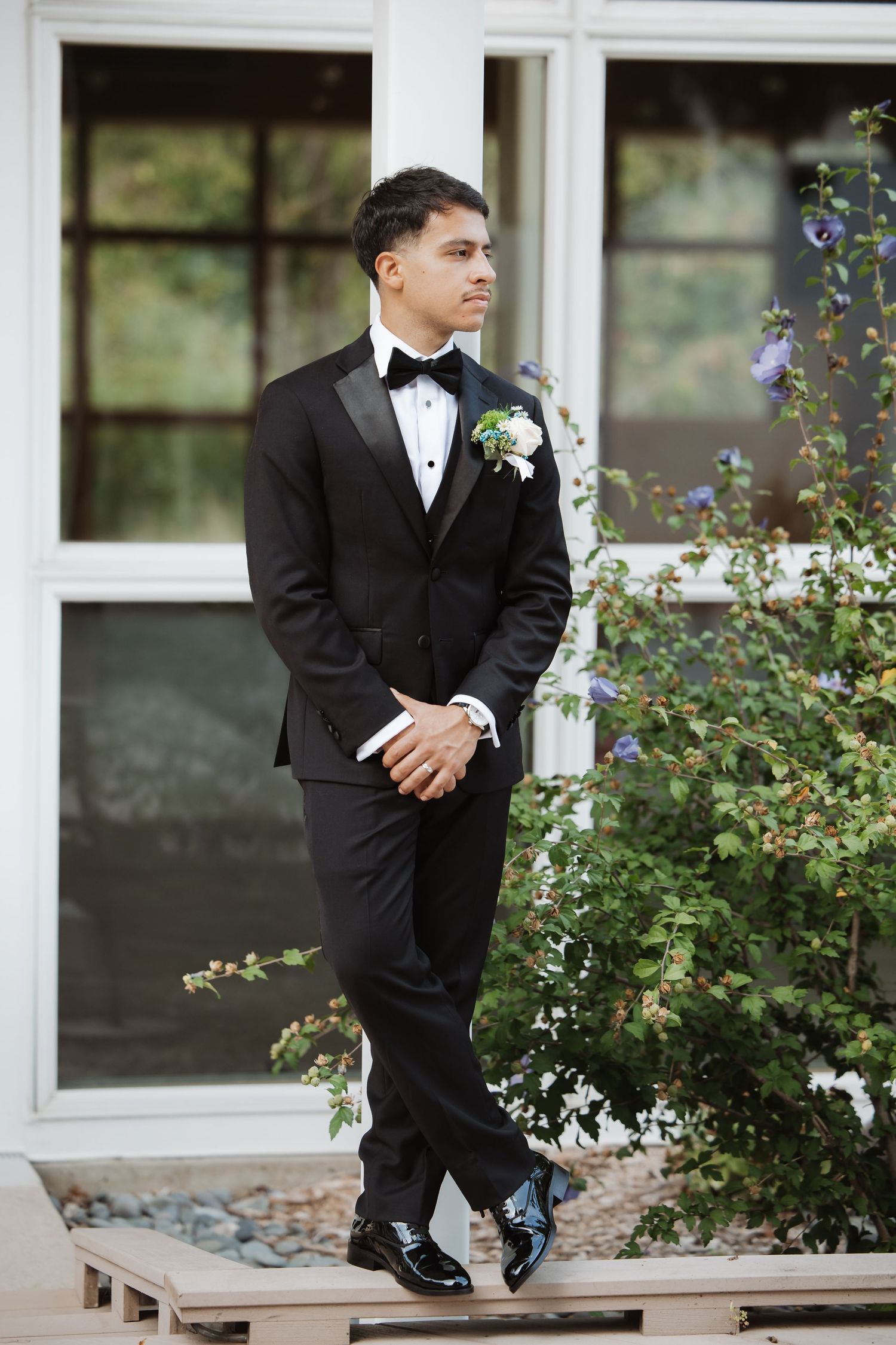 Elegant groom in black tuxedo and bow tie poses near blooming blue flowers.