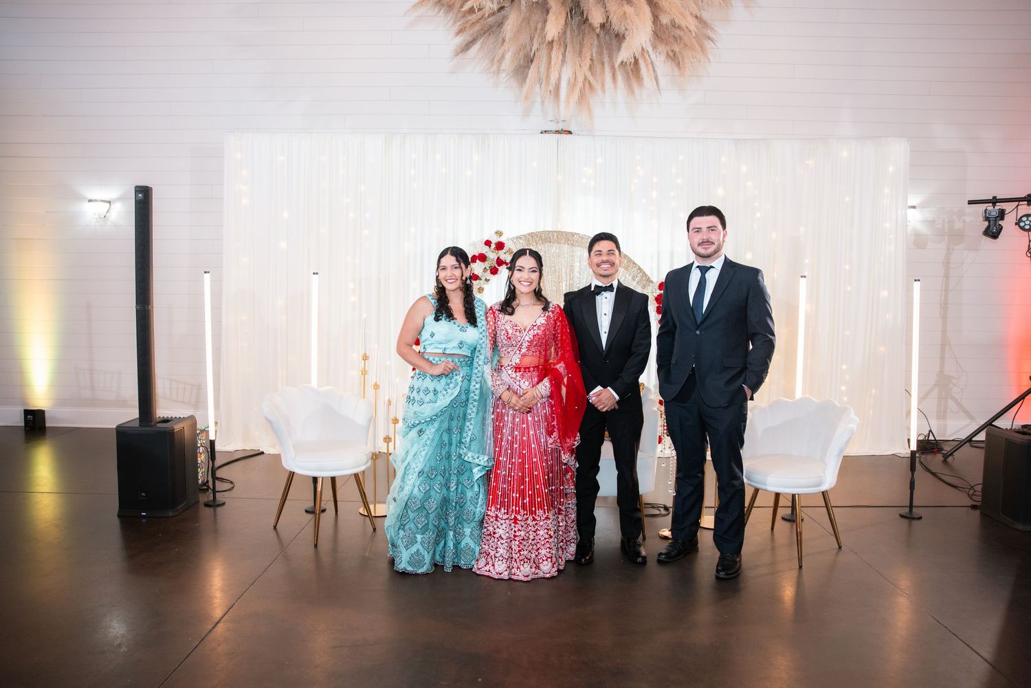 Wedding couple in traditional attire poses with two guests in front of elegant white backdrop.