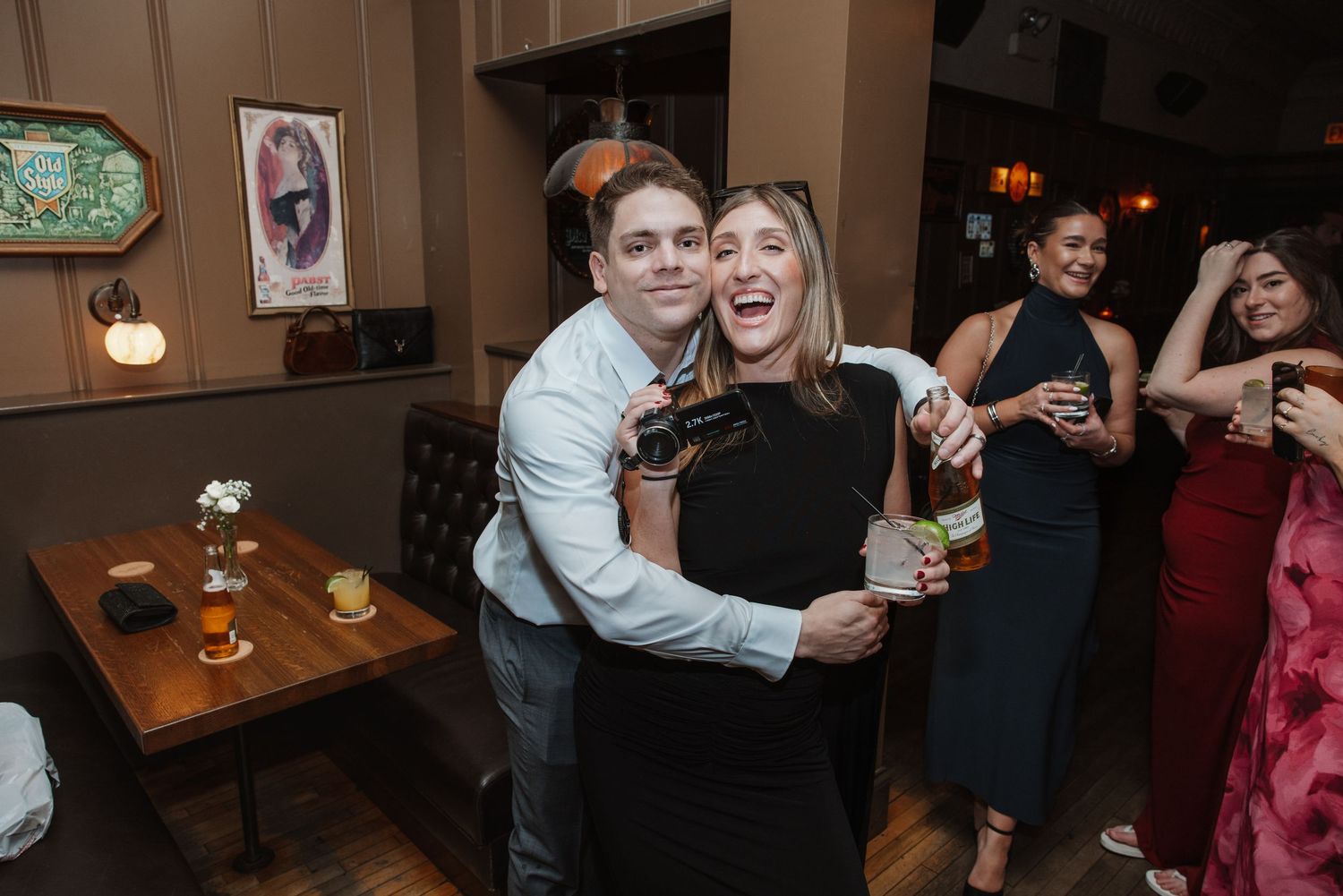 Man hugs laughing woman holding drink at lively indoor wedding reception party.