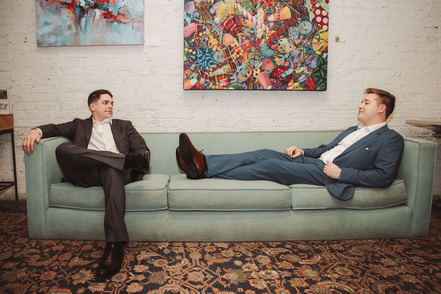 Two men in suits sit on a mint green mid-century sofa beneath colorful abstract paintings in a stylish loft.