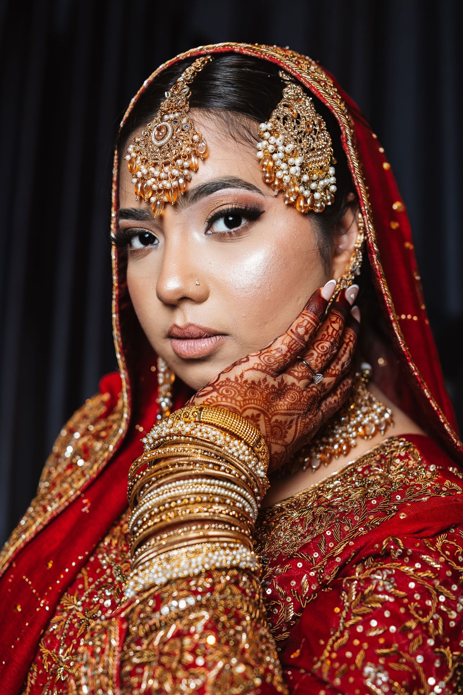 Indian bride in red lehenga with gold jewelry, maang tikka, bangles, and intricate henna on hands.