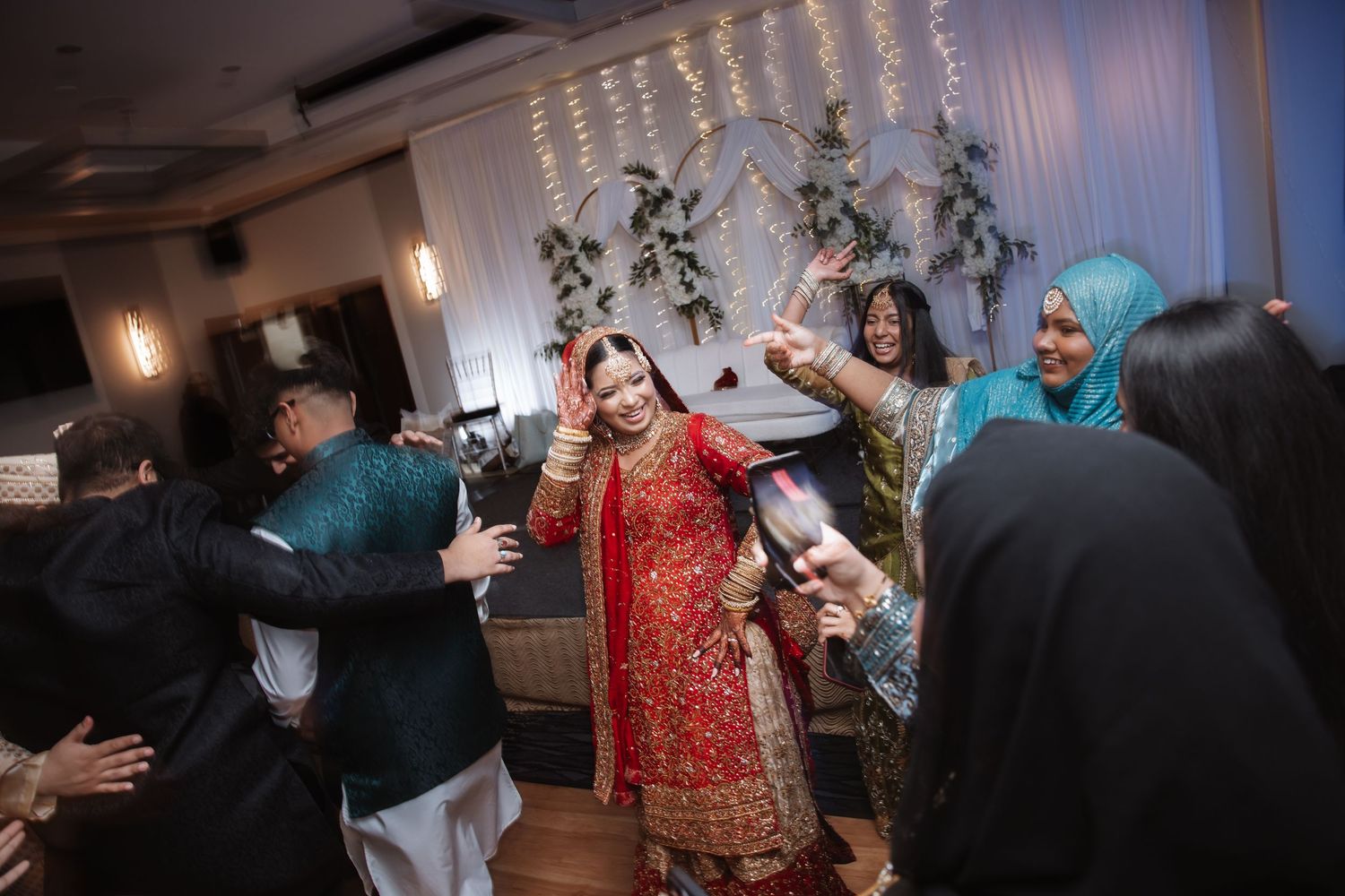 South Asian bride in red lehenga dances joyfully surrounded by guests at wedding reception.